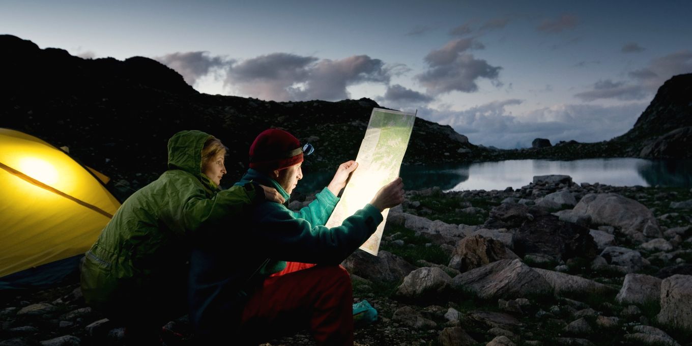 Two campers read a map by a tent on a rocky lake shore at dusk